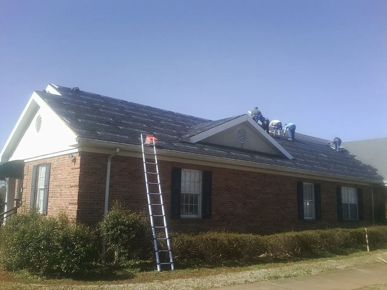 Crew tearing shingles off a brick ranch roof with a ladder set — Metal Roof Repair work by our crew in Eastman