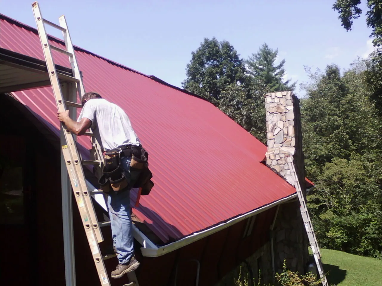 Roofer on a ladder beside a red metal roof and stone chimney in Eastman